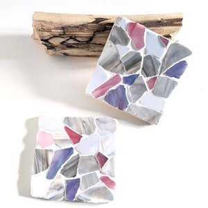 Puede incluir: Dos posavasos cuadrados hechos de azulejos de mosaico. Los azulejos son en tonos de gris, blanco, rosa y morado. Los posavasos están sobre un trozo de madera flotante.