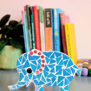 May include: A mosaic elephant figurine in shades of blue, white, and red. The elephant has a black eye and is decorated with triangular tiles. Books of various colors are in the background.