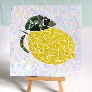 May include: A mosaic artwork of a lemon on a white background. The lemon is made of small, yellow, green, and white tiles. The artwork is displayed on a small wooden easel.