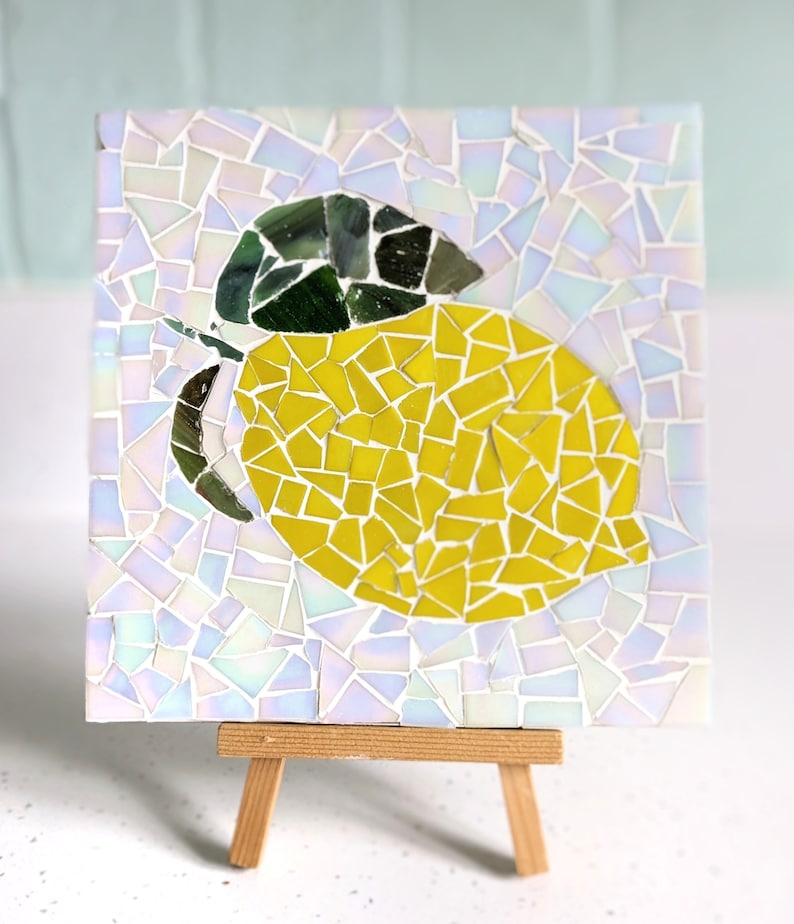 May include: A mosaic artwork of a lemon on a white background. The lemon is made of small, yellow, green, and white tiles. The artwork is displayed on a small wooden easel.