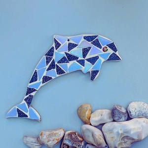 Può includere: Un delfino a mosaico nei toni del blu e del bianco. Il delfino è composto da tessere triangolari e ha un piccolo occhio rosso. Una collezione di pietre lisce e arrotondate è disposta sotto il delfino su uno sfondo azzurro.