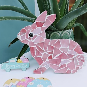 Può includere: Una statuetta di coniglio a mosaico rosa con un occhio nero e una coda bianca. Il coniglio è fatto di varie tonalità di pezzi di vetro rosa. Un piccolo camioncino azzurro con la scritta "Happy Easter" è accanto al coniglio.
