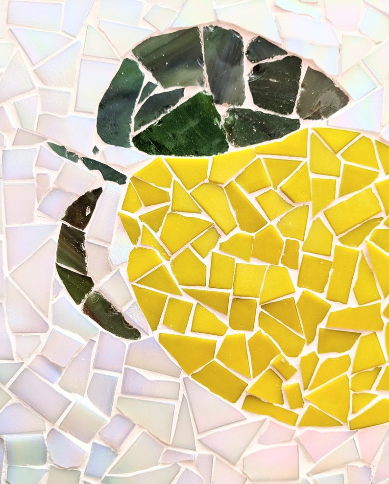 May include: A mosaic artwork depicting a lemon with a green leaf. The lemon is made of small yellow tiles, and the leaf is made of small green tiles. The background is made of small white tiles.