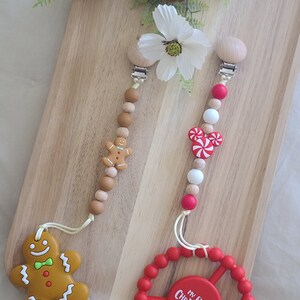 May include: Two Christmas themed baby teethers with wooden beads and clips. One teether features a gingerbread man and the other features a candy cane and a Mickey Mouse head. The third teether is a red silicone ring with the text "My First Christmas!"