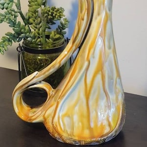 May include: A ceramic pitcher with a unique, abstract design. The pitcher is a light yellow color with white and brown streaks. It has a long, curved spout and a handle.