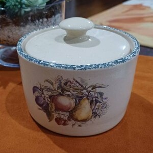 May include: A white ceramic canister with a lid, featuring a floral design with fruit, including a pear, apple, and plums. The canister has a green and blue trim around the top.