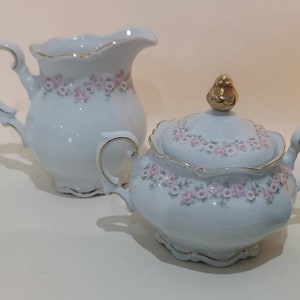 May include: A white porcelain creamer and sugar bowl set with a delicate floral pattern in pink and gold accents. The creamer has a handle and a spout, while the sugar bowl has a lid with a gold bird finial.