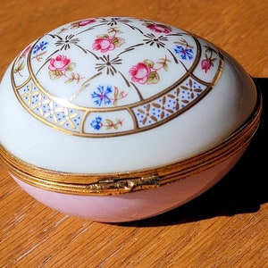May include: An egg-shaped trinket box with a hinged lid. The top is white with a floral design in pink, blue, and gold. The bottom is light pink, and the box is trimmed with gold. The box is on a wooden surface.