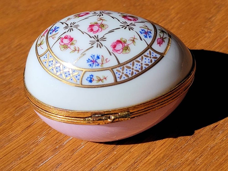 May include: An egg-shaped trinket box with a hinged lid. The top is white with a floral design in pink, blue, and gold. The bottom is light pink, and the box is trimmed with gold. The box is on a wooden surface.