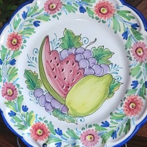 May include: A white ceramic plate with a blue rim and a hand-painted design of fruit, including a watermelon, lemon, and grapes. The plate is decorated with pink and blue flowers and green leaves.