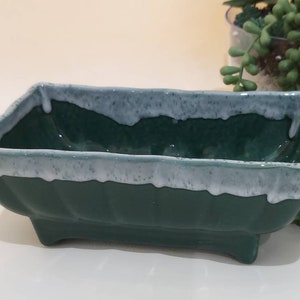 May include: A rectangular green ceramic planter with a white glaze around the rim. The planter has a slightly flared lip and four small feet.