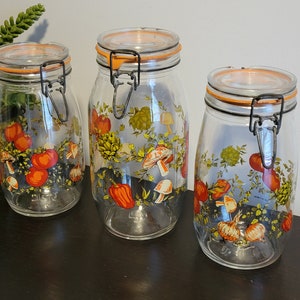May include: Set of three glass storage jars with orange and green floral designs featuring mushrooms, tomatoes, peppers, and onions. The jars have metal lids with a locking mechanism.