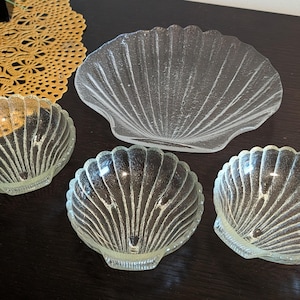 May include: A set of four clear glass dishes shaped like seashells, featuring a textured surface with radiating ridges. The collection includes one large shallow platter and three smaller, deeper bowls, all crafted from transparent glass material.