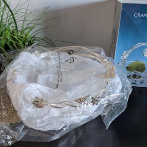 May include: Silver-colored metal fruit basket with a grape design, still in its original packaging. The basket has a handle and is partially visible through the clear plastic. The box in the background reads "Silver Fruit Grape Design Basket."