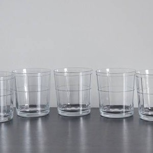 May include: Six clear glass tumblers with a textured design. The glasses are arranged in a row on a dark surface.