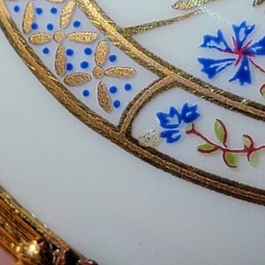 May include: Close-up of a white, ornate object with gold trim and floral designs. The gold trim features a repeating pattern of gold flowers and blue dots. Blue and red floral accents are also visible.