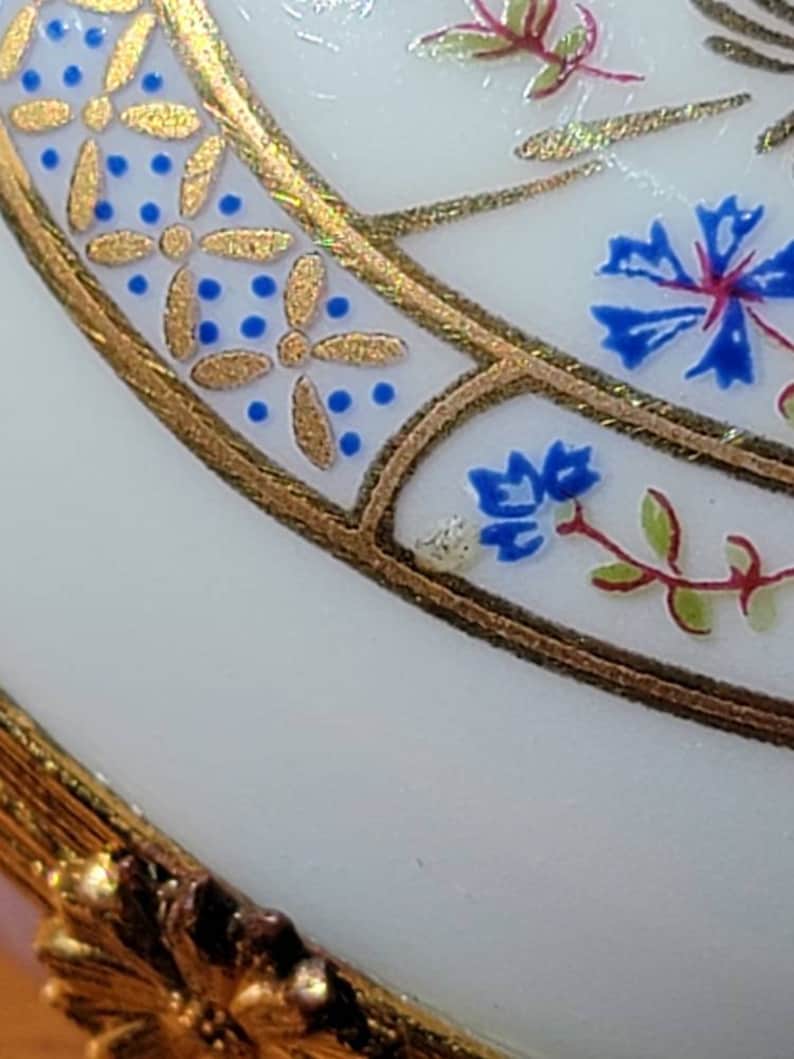 May include: Close-up of a white, ornate object with gold trim and floral designs. The gold trim features a repeating pattern of gold flowers and blue dots. Blue and red floral accents are also visible.