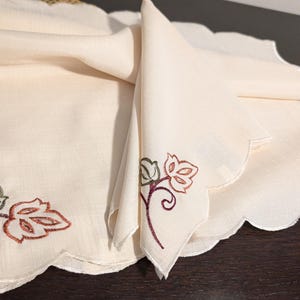 May include: Cream-colored linen table runner and matching napkin set. The runner and napkin have a scalloped edge and embroidered leaf designs in shades of brown, green, and burgundy. The set is folded on a dark wood surface.