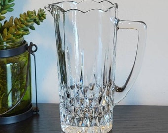 May include: A clear glass pitcher with a scalloped rim and a diamond-shaped pattern. The pitcher has a handle and is perfect for serving beverages.