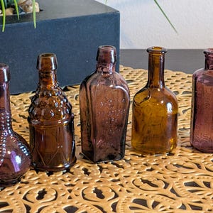May include: Five antique glass bottles in shades of purple and amber are arranged on a decorative surface. The bottles vary in shape and design, showcasing embossed patterns and unique forms. The collection offers a vintage aesthetic.