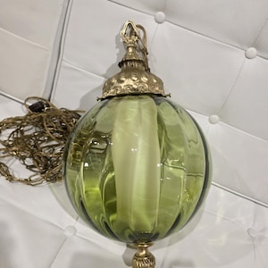 May include: A spherical pendant light fixture featuring a translucent olive green glass shade with vertical ribbed detailing. The lamp includes an ornate brass-toned metal cap at the top and a matching decorative finial at the bottom, suspended by a chain.