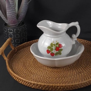 May include: A white ceramic pitcher and bowl set with a strawberry design. The pitcher has a handle and a wide mouth. The bowl is round and has a scalloped edge. The set is on a woven tray.