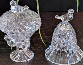 May include: Two clear glass decorative pieces. One is a pedestal dish with a lid, topped with a small bird. The other is a bell-shaped piece, also with a bird on top. Both have a diamond pattern.