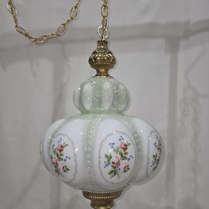 May include: A hanging pendant lamp featuring a milk glass shade with a pale green tint and a bulbous, paneled shape. The glass is decorated with floral decals of pink roses and blue flowers. It includes a gold-tone metal chain and hardware.