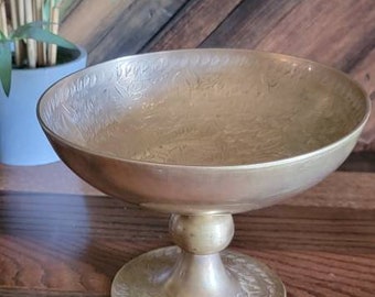 May include: A gold-toned metal bowl with an ornate design sits on a pedestal. The bowl has a wide, shallow shape and is decorated with intricate floral patterns.