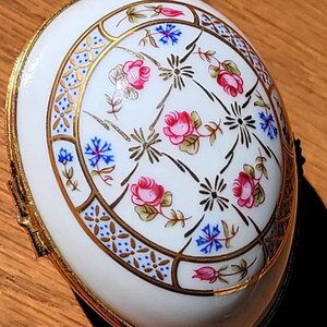 May include: White, oval-shaped trinket box with a gold-colored trim. The lid features a floral design with pink roses, blue flowers, and gold accents. The box is likely made of porcelain or a similar material.