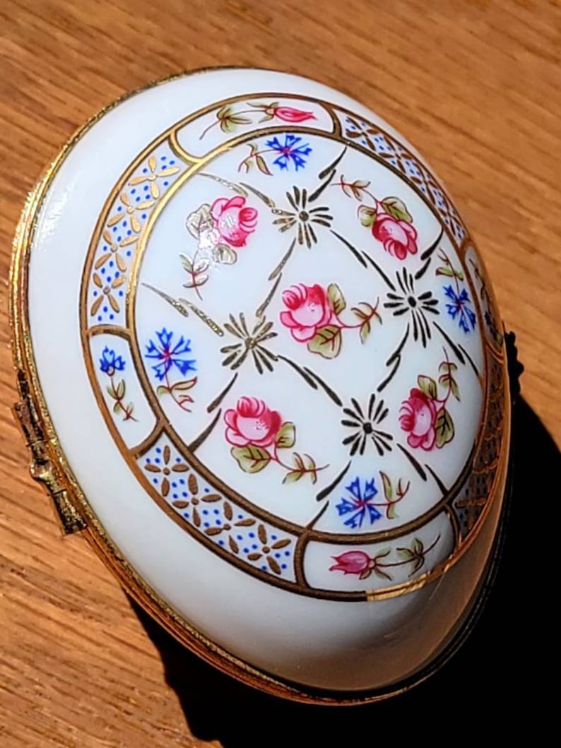 May include: White, oval-shaped trinket box with a gold-colored trim. The lid features a floral design with pink roses, blue flowers, and gold accents. The box is likely made of porcelain or a similar material.