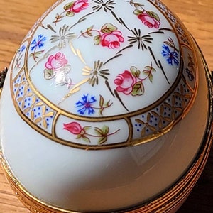 May include: A white, egg-shaped decorative container with a hinged lid. The container is adorned with pink roses, blue flowers, and gold accents. The base and rim are trimmed with gold. The design is reminiscent of fine porcelain.