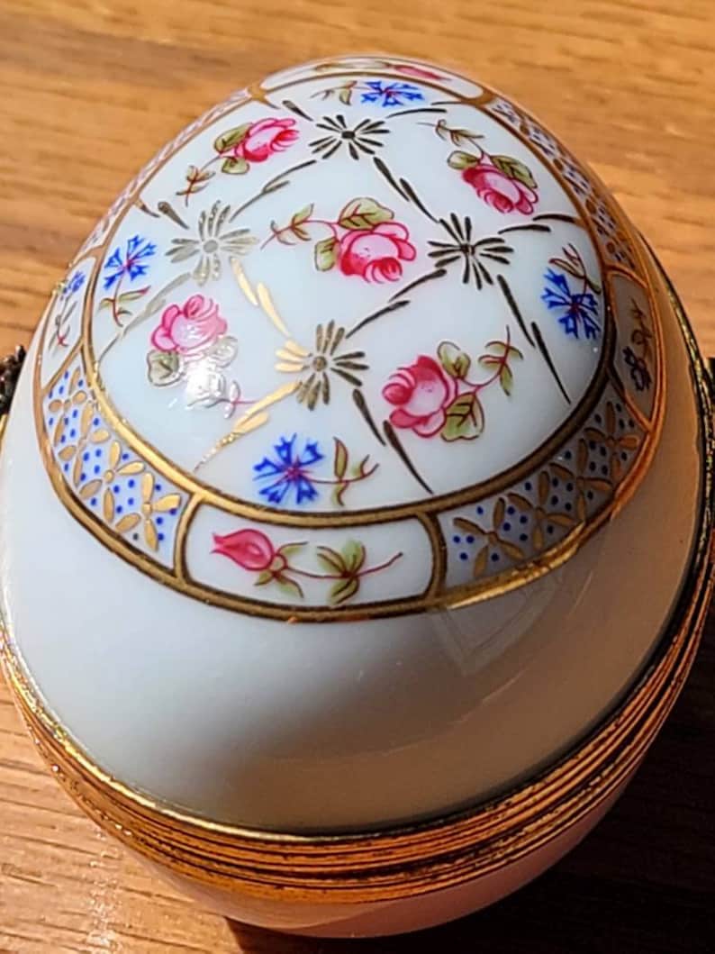 May include: A white, egg-shaped decorative container with a hinged lid. The container is adorned with pink roses, blue flowers, and gold accents. The base and rim are trimmed with gold. The design is reminiscent of fine porcelain.