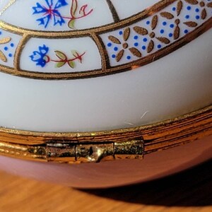 May include: Close-up of a decorative trinket box. The box is primarily white with a pink base and gold trim. The lid features a floral design with blue and gold accents. The box is hinged and appears to be made of porcelain.