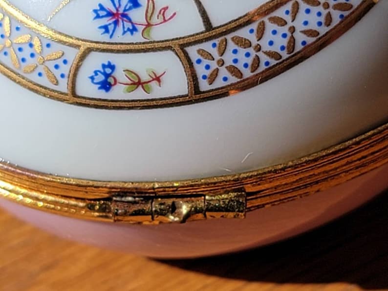 May include: Close-up of a decorative trinket box. The box is primarily white with a pink base and gold trim. The lid features a floral design with blue and gold accents. The box is hinged and appears to be made of porcelain.