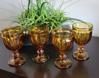 May include: Four amber-colored glass wine goblets with a textured design. The goblets are arranged on a dark brown surface.