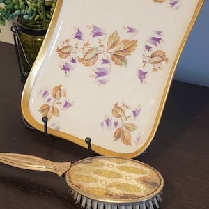 May include: A white ceramic tray with a gold rim and a floral design featuring purple flowers and brown leaves. The tray is on a black stand. A gold and brown hairbrush is on a dark wood surface.