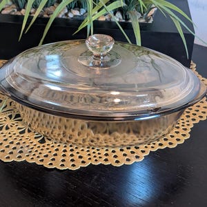May include: Oval-shaped, clear glass casserole dish with a matching lid and a clear knob. The dish has a textured, hammered appearance. The lid has a decorative etched design. The dish sits on a gold-colored, patterned placemat.