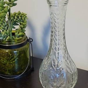 May include: A clear glass vase with a textured leaf pattern. The vase has a tall, slender neck and a rounded base.