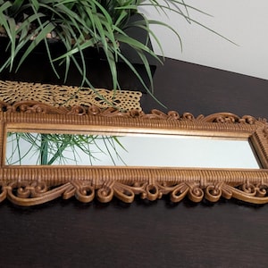 May include: A rectangular, ornate, gold-colored mirror with a reflective surface. The frame features intricate scrollwork and a textured border. The mirror is a decorative home accent.