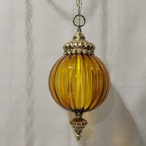 May include: A vintage amber glass pendant light with a ribbed, globe-shaped shade. The light fixture has ornate brass-colored metal accents and a chain for hanging. The overall design is reminiscent of mid-century modern decor.