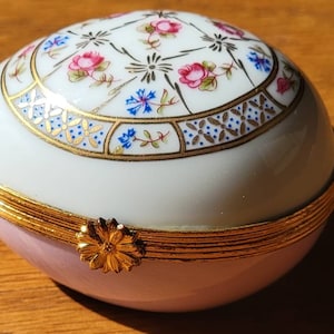 May include: A decorative, oval-shaped trinket box. The top is white with a floral design in pink, blue, and gold. The bottom is light pink, and the box is trimmed with gold. A gold flower clasp secures the lid.