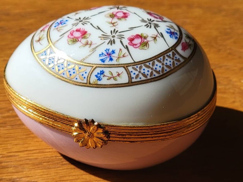May include: A decorative, oval-shaped trinket box. The top is white with a floral design in pink, blue, and gold. The bottom is light pink, and the box is trimmed with gold. A gold flower clasp secures the lid.