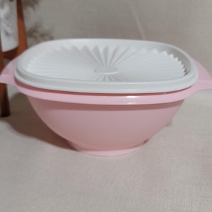 May include: A pink plastic bowl with a white lid. The bowl has a rounded shape with a slightly flared rim and two small handles. The lid is white with a textured, fan-like design.