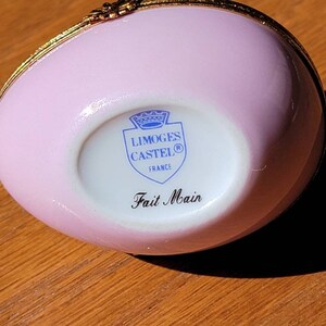 May include: A pink, egg-shaped Limoges Castel trinket box with gold trim. The bottom of the box features the Limoges Castel logo and the words "Fait Main". The box is made in France.