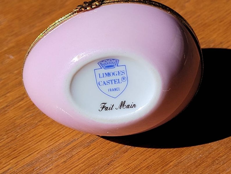 May include: A pink, egg-shaped Limoges Castel trinket box with gold trim. The bottom of the box features the Limoges Castel logo and the words "Fait Main". The box is made in France.
