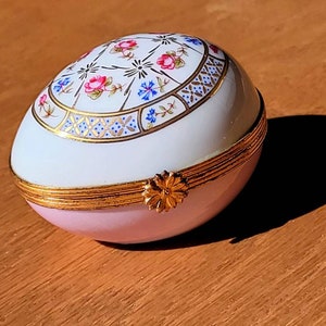 May include: An egg-shaped trinket box with a hinged lid. The box is primarily pink and white, with a gold-colored band around the middle and a floral design on the top. A small gold-colored flower serves as a clasp.