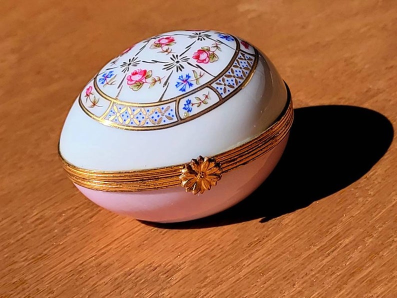 May include: An egg-shaped trinket box with a hinged lid. The box is primarily pink and white, with a gold-colored band around the middle and a floral design on the top. A small gold-colored flower serves as a clasp.