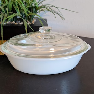 May include: A white oval casserole dish with a clear glass lid. The lid has a small, clear knob handle. The dish is sitting on a dark surface, with a plant in the background.