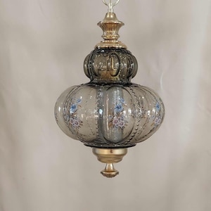 May include: A vintage hanging light fixture with a smoky gray glass shade. The shade is adorned with delicate floral patterns in blue and pink. The fixture has gold-colored metal accents and a chain for hanging.